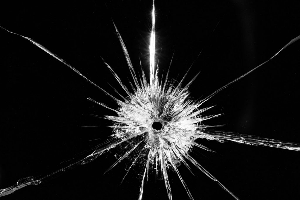 Close up of broken, cracked pane of glass with central bullet hole, on black background